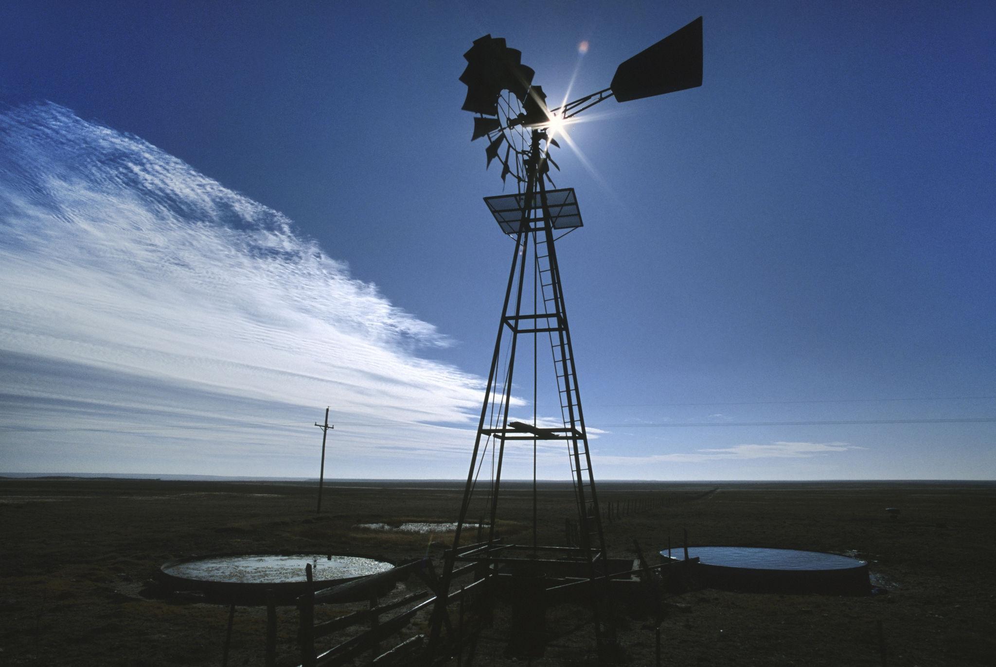 A silhouetted windmill stands against a blue sky, with sunlight peeking through its blades. Nearby, a circular water trough reflects the landscape.